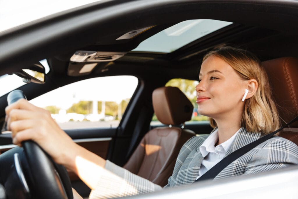 Mulher jovem loira sorrindo dirige carro com teto solar, usando fone wireless branco e blazer xadrez cinza