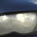 Mão apontando para farol de carro embaçado com lente opaca e sinais de umidade interna