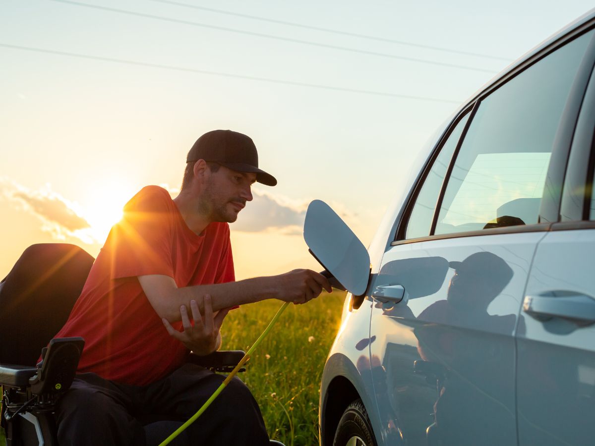 Homem em cadeira de rodas conecta carregador a carro branco ao pôr do sol em campo verde