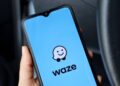 Celular com aplicativo Waze exibindo logo e mascote em tela azul, segurado por mão com unhas claras dentro de carro