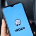Celular com aplicativo Waze exibindo logo e mascote em tela azul, segurado por mão com unhas claras dentro de carro