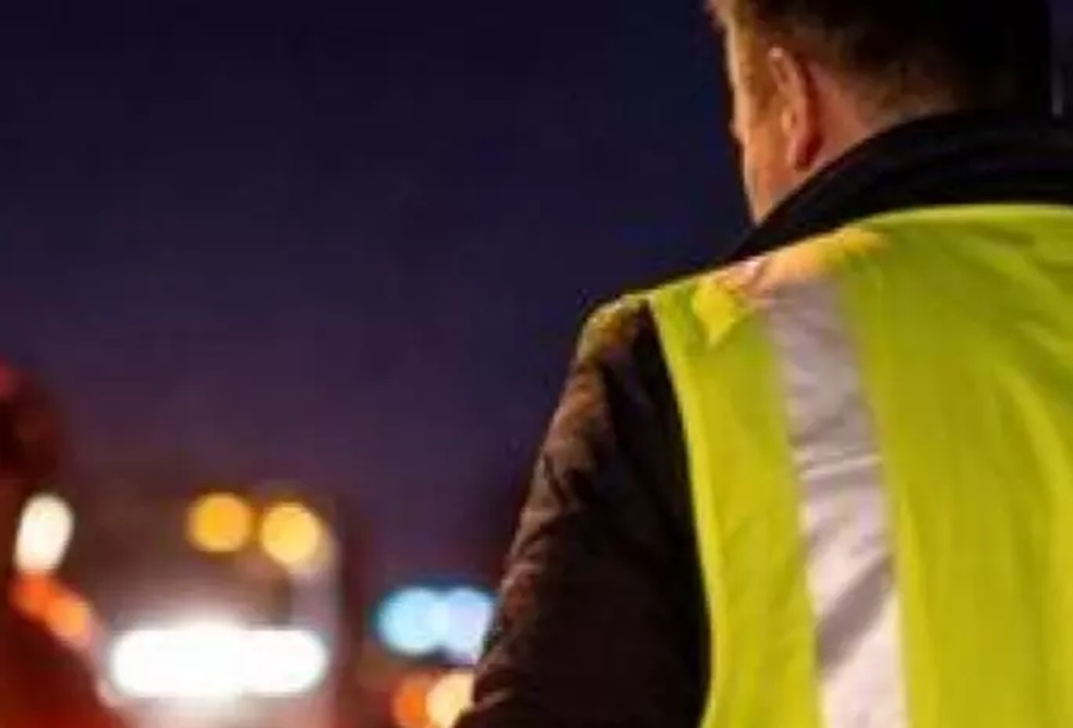 Homem de costas usa colete amarelo fluorescente com faixa cinza, observando luzes de veículos desfocadas à noite