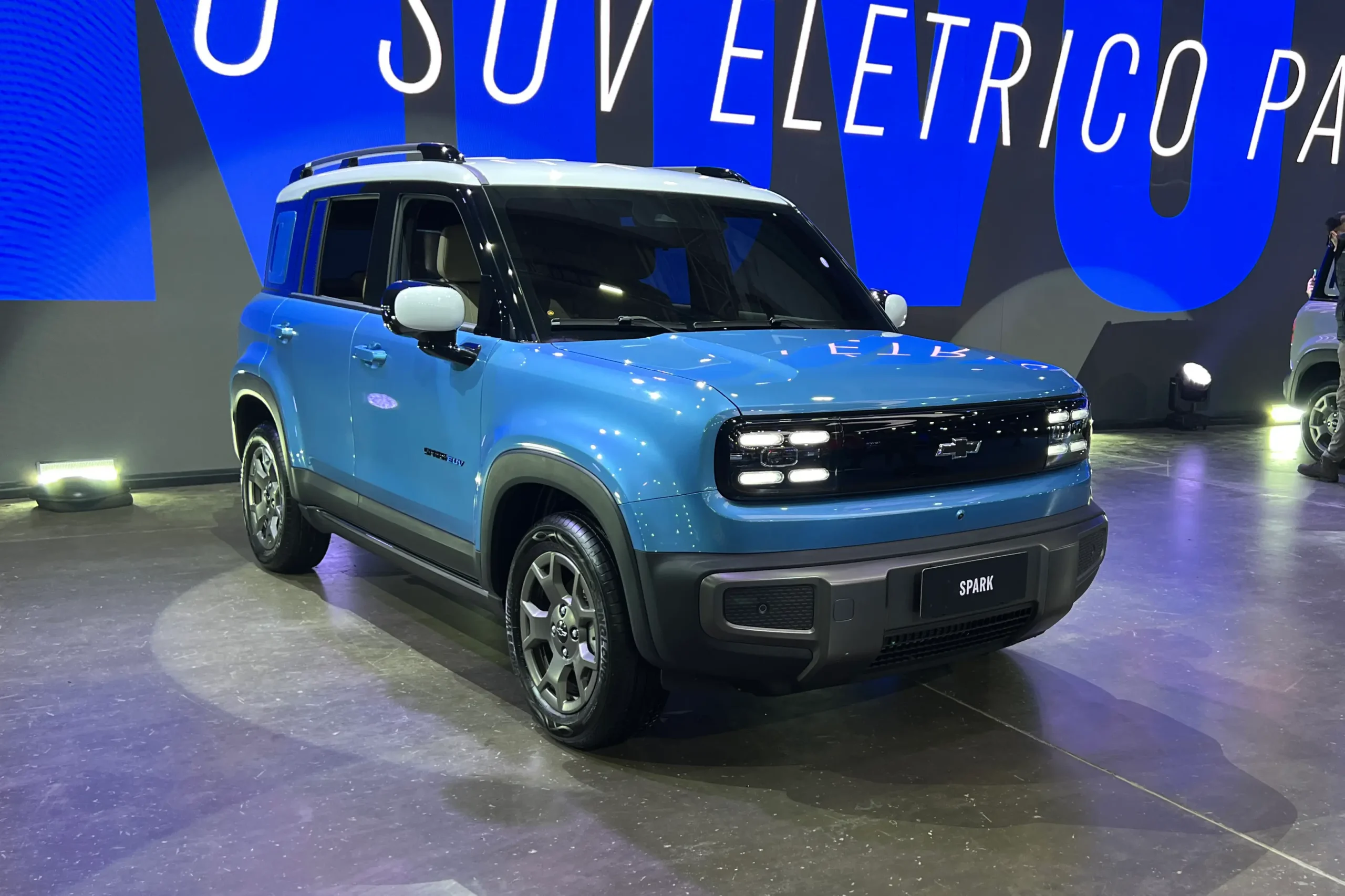 SUV elétrico Chevrolet Spark EUV azul com teto branco, exposto em palco iluminado com painel azul ao fundo