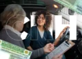 Mulher de cabelos cacheados ao volante recebe instruções de examinador com prancheta, letras CNH sobrepostas