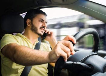 Homem de barba, camisa amarela, fala ao celular segurando o aparelho na orelha enquanto dirige o veículo.