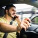 Homem de barba, camisa amarela, fala ao celular segurando o aparelho na orelha enquanto dirige o veículo.
