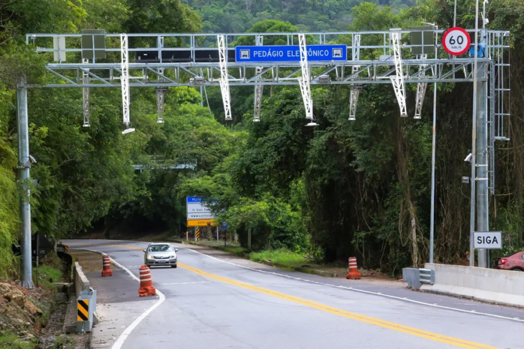 Carro passando sob pórtico de pedágio eletrônico com placa "PEDÁGIO ELETRÔNICO" em rodovia cercada por vegetação.