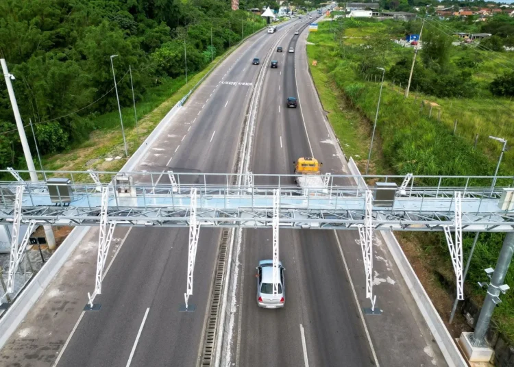Rodovia de pista dupla com pórtico metálico de pedágio eletrônico free flow, veículos trafegando e vegetação ao redor.