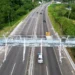 Rodovia de pista dupla com pórtico metálico de pedágio eletrônico free flow, veículos trafegando e vegetação ao redor.