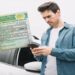 Homem jovem usa celular ao lado de carro branco enquanto mão segura carteira de motorista brasileira