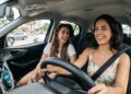 Duas mulheres jovens, sorrindo, dirigem e conversam em carro moderno, trânsito ao fundo