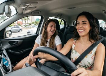 Duas mulheres jovens, sorrindo, dirigem e conversam em carro moderno, trânsito ao fundo