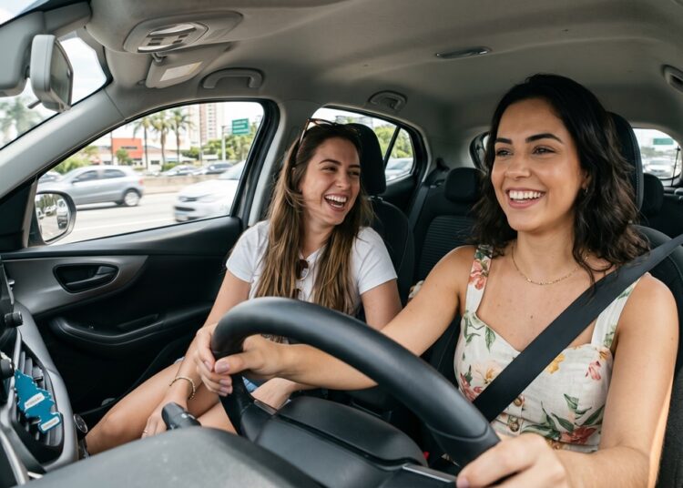 Duas mulheres jovens, sorrindo, dirigem e conversam em carro moderno, trânsito ao fundo