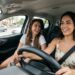 Duas mulheres jovens, sorrindo, dirigem e conversam em carro moderno, trânsito ao fundo