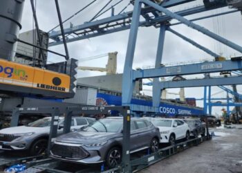 Carros elétricos cinza e branco sobre plataforma no porto, guindaste Login, navio Cosco Shipping ao fundo