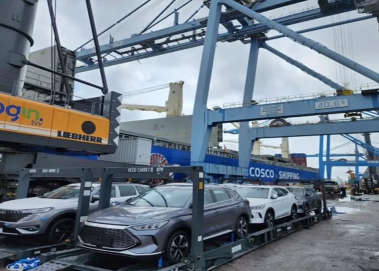 Carros elétricos cinza e branco sobre plataforma no porto, guindaste Login, navio Cosco Shipping ao fundo