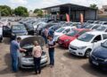 Grupo de pessoas avaliando carros em pátio de loja de usados com preços nos para-brisas
