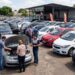 Grupo de pessoas avaliando carros em pátio de loja de usados com preços nos para-brisas