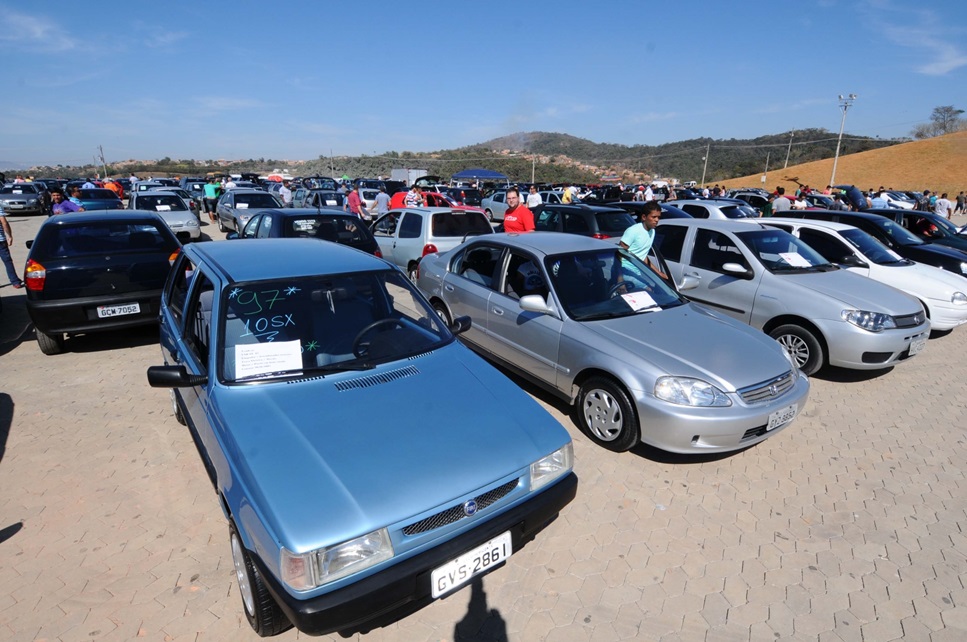 Vários carros usados expostos em pátio ao ar livre, pessoas observando veículos marcados com anos e placas