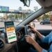 Mulher dirige carro se aproximando de pedágio, celular mostra alerta do aplicativo CNH Digital sobre tarifa sem parar