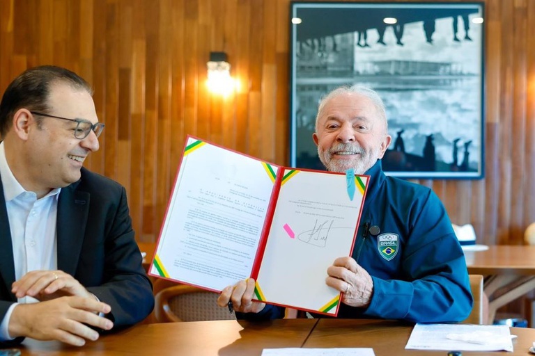 Presidente Lula exibe documento assinado ao lado de homem em sala com parede de madeira