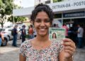 Jovem sorrindo mostra CNH provisória em frente ao Detran-BA, placa de atendimento ao fundo