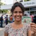 Jovem sorrindo mostra CNH provisória em frente ao Detran-BA, placa de atendimento ao fundo