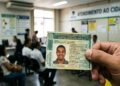 Mão segura CNH brasileira em sala de atendimento ao cidadão, pessoas sentadas aguardam