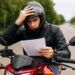 Homem jovem de capacete preto lê papel na moto vermelha com expressão preocupada em rua arborizada