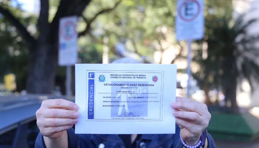 Pessoa segura credencial de estacionamento para vaga reservada em via pública com placas ao fundo