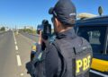 Agente da PRF com colete tático e boné aponta radar portátil em rodovia, viatura azul ao lado, céu azul ao fundo