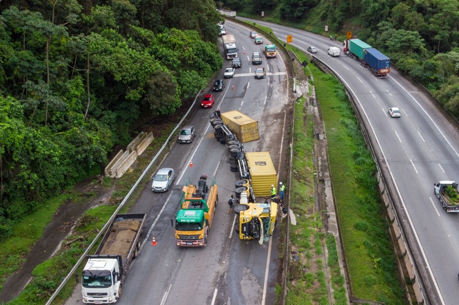 Vista aérea de carreta amarela tombada com contêineres na pista de rodovia cercada por mata verde, equipes de resgate e fila de veículos