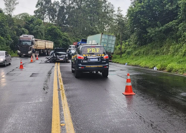 Viatura da PRF com cones laranjas em rodovia molhada de pista simples, caminhões e carro acidentado ao fundo