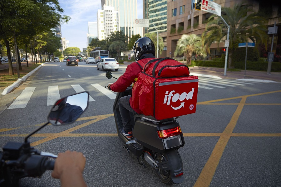 Motociclista com jaqueta vermelha e capacete preto pilota moto com bag térmica vermelha do iFood em avenida urbana