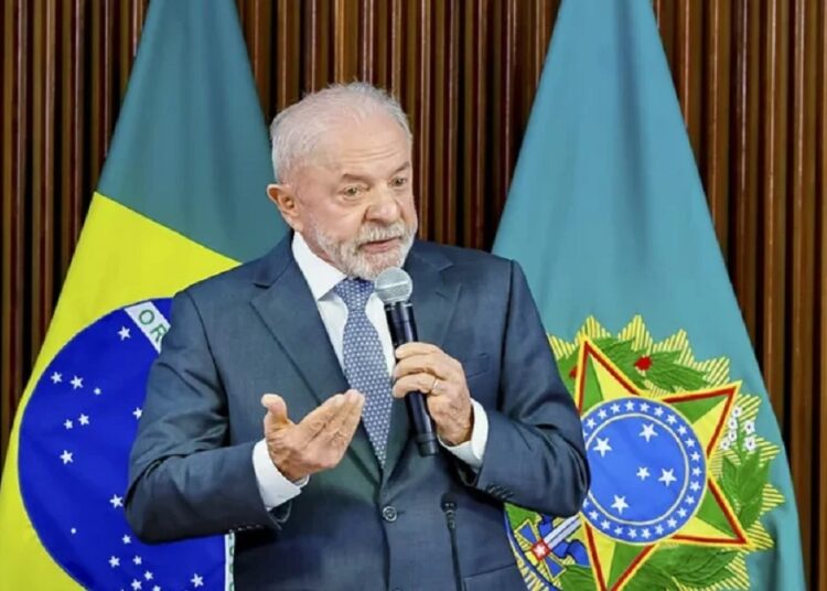 Presidente Lula discursa com microfone, terno azul e gravata clara, diante das bandeiras do Brasil e presidencial