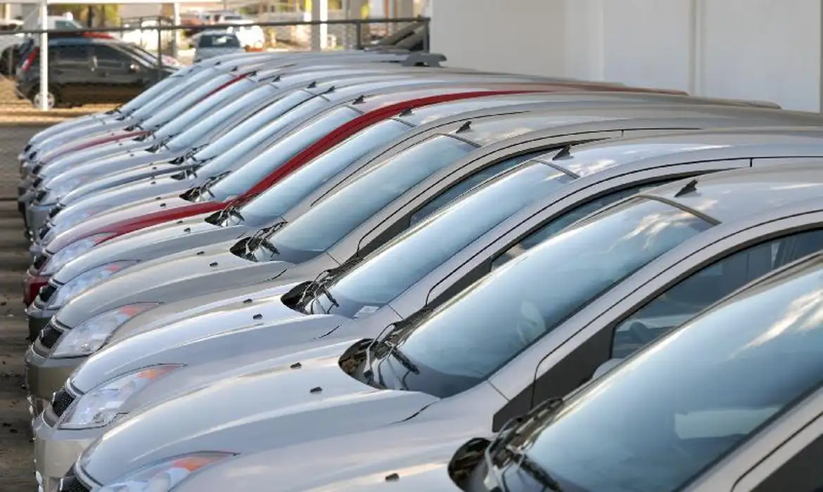 Vários carros prateados e um vermelho alinhados em fila em pátio externo de concessionária