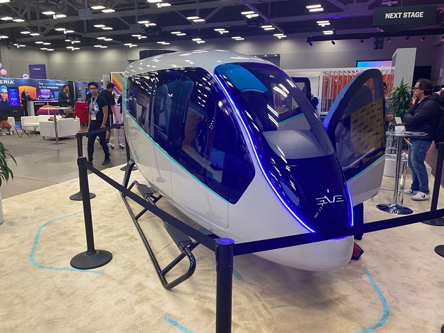 Cabine futurista branca e azul do Eve 100 com iluminação LED azul, portas abertas e logo EVE visível, em pavilhão de feira