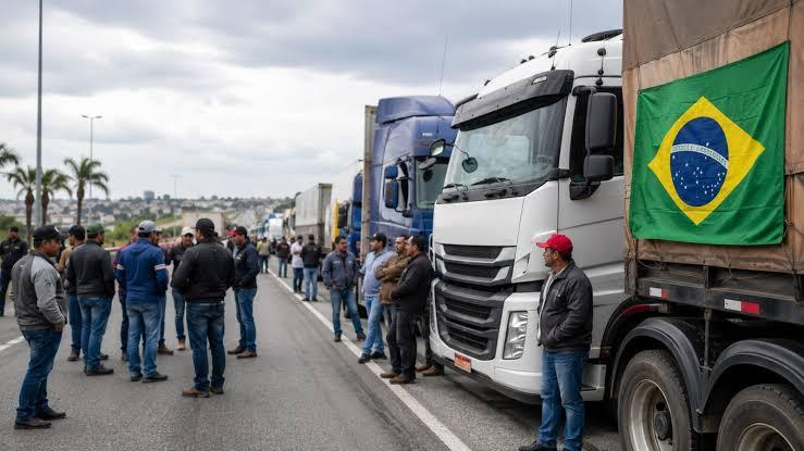 Caminhoneiros reunidos ao lado de caminhões estacionados na rodovia, bandeira do Brasil em destaque, céu nublado