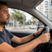 Homem adulto dirigindo carro, olhando à frente, com celular usando GPS no painel, cidade ao fundo