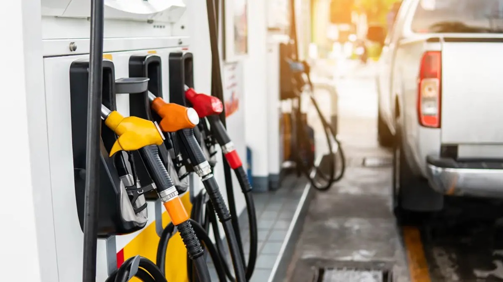Bicos de abastecimento amarelo, laranja e vermelho em bomba de posto de gasolina, com veículo prata ao fundo