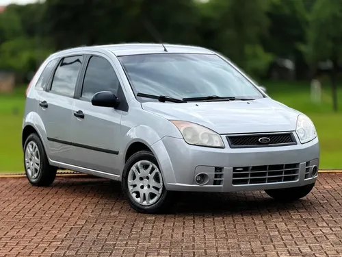 Ford Fiesta prata estacionado, vidro escuro, fundo verde e chão de tijolos