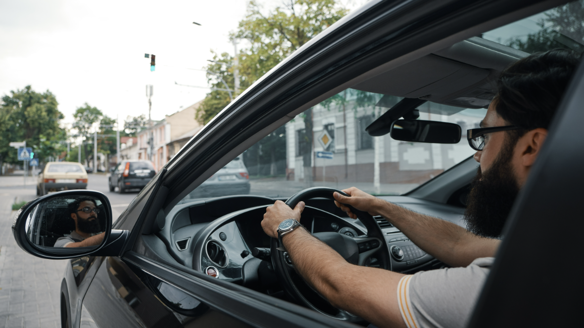 Motorista de barba dirige carro preto em rua urbana.