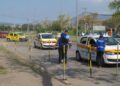 Carros de autoescola do Detran-RJ em teste prático, avaliadores em coletes azuis interagem com candidatos na pista delimitada