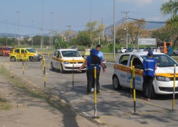 Carros de autoescola do Detran-RJ em teste prático, avaliadores em coletes azuis interagem com candidatos na pista delimitada