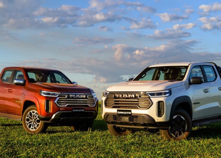 Duas picapes Ram Dakota 2026, uma laranja Laramie e outra branca Warlock, estacionadas em gramado verde sob céu azul