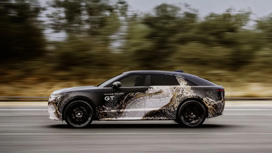 Protótipo Range Rover GT camuflado com padrão dourado em movimento na estrada, exibindo silhueta de sedã