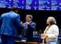 Três senadores interagem no plenário do Senado; homem ao centro faz sinal de positivo, ao lado de mulher de cabelos grisalhos, simbolizando a MP do frete.