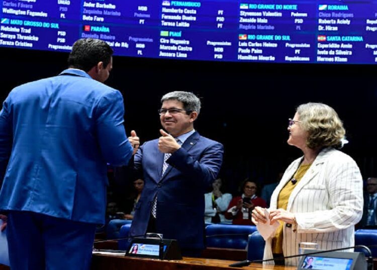 Três senadores interagem no plenário do Senado; homem ao centro faz sinal de positivo, ao lado de mulher de cabelos grisalhos, simbolizando a MP do frete.
