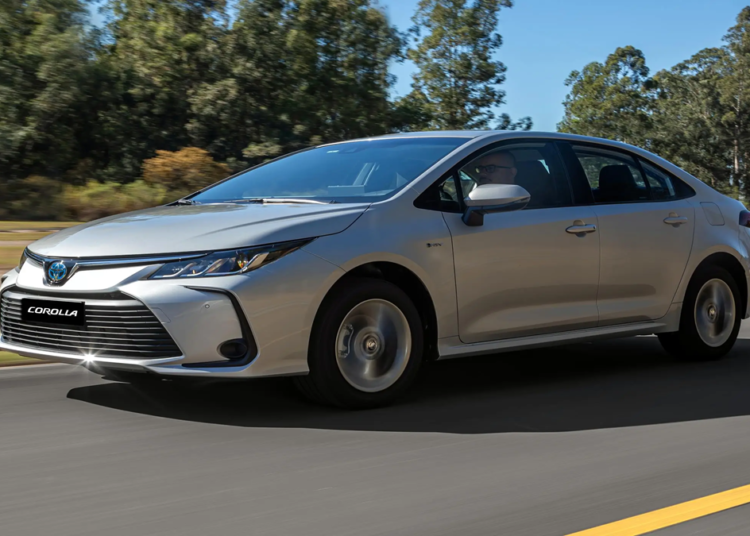 Toyota Corolla sedan prata trafega em rodovia com fundo de vegetação verde, exibindo grade frontal com emblema da marca