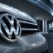 Emblema cromado da Volkswagen em close na parte frontal de carro prateado, fundo urbano desfocado